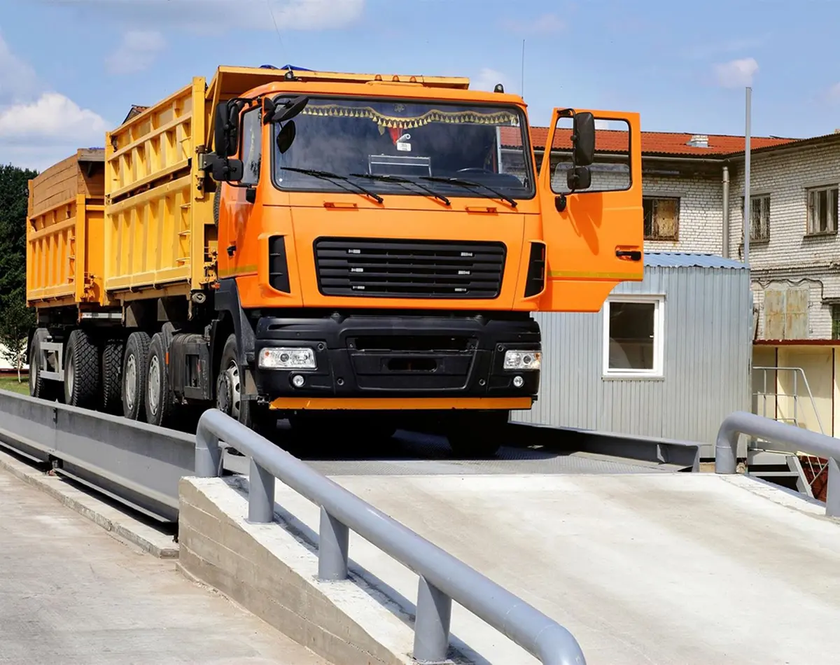 Large orange dump truck parked on an above-ground industrial weighbridge platform with a scale house and site office in the background