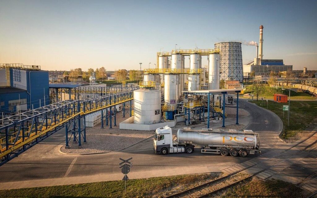 Tanker truck driving past a large industrial chemical processing plant with vertical storage silos, cooling towers, and overhead piping networks